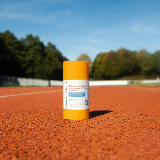 Le baume chauffant 100% naturel et rechargeable IZOM Athlétique se situe sur une piste d'athlétisme. Le stick est orange avec une étiquette blanche. La contenance est de 70g pour une durée d'utilisation de 5 à 6 mois. Ce baume permet d'apaiser les tensions musculaires et articulaires.