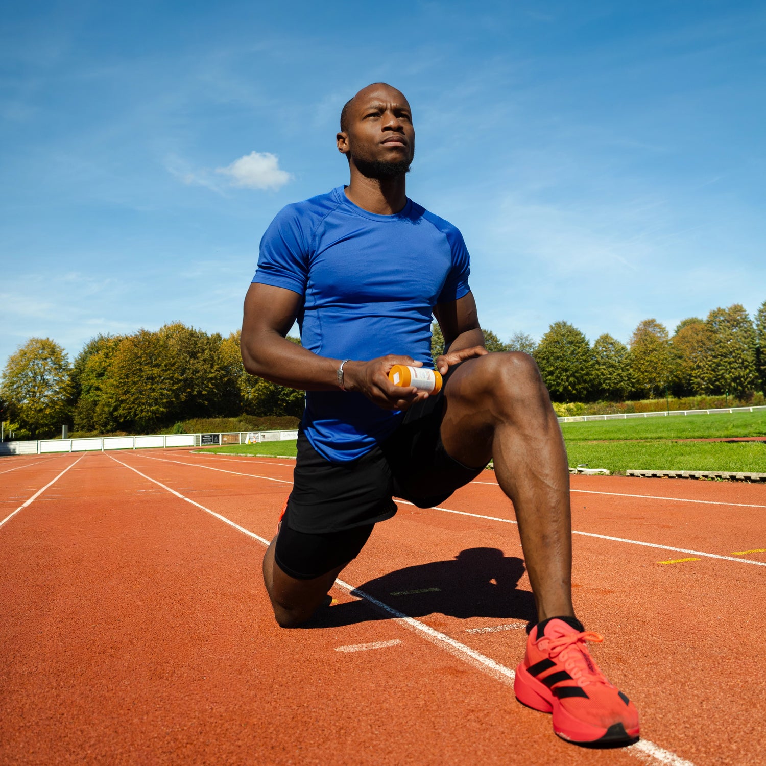 Oumar BAMBA, Sprinteur et client d'IZOM Athlétique applique le baume chauffant 100% naturel et rechargeable IZOM Athlétique sur sa cuisse. Il se situe sur une piste d'athlétisme orange. Le format du stick est de 70g pour une durée d'utilisation de 5 à 6 mois.