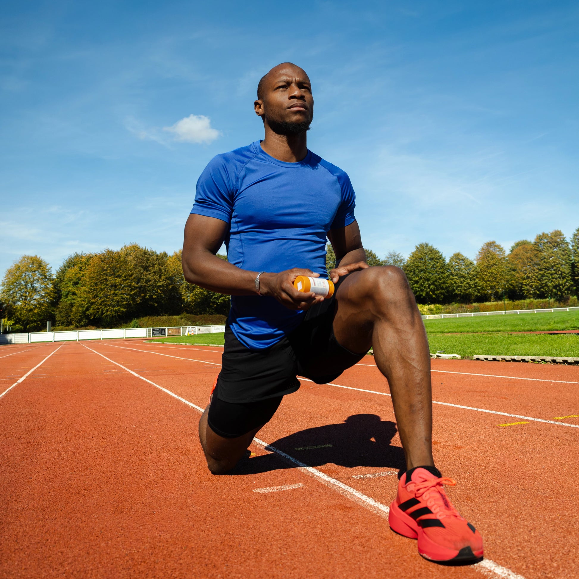 Oumar BAMBA, Sprinteur et client d'IZOM Athlétique applique le baume chauffant 100% naturel et rechargeable IZOM Athlétique sur sa cuisse. Il se situe sur une piste d'athlétisme orange. Le format du stick est de 70g pour une durée d'utilisation de 5 à 6 mois.