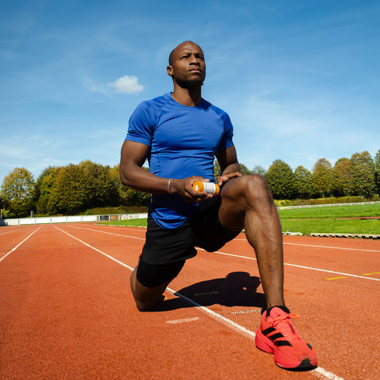 Oumar BAMBA, Sprinteur et client d'IZOM Athlétique applique le baume chauffant 100% naturel et rechargeable IZOM Athlétique sur sa cuisse. Il se situe sur une piste d'athlétisme orange. Le format du stick est de 70g pour une durée d'utilisation de 5 à 6 mois.