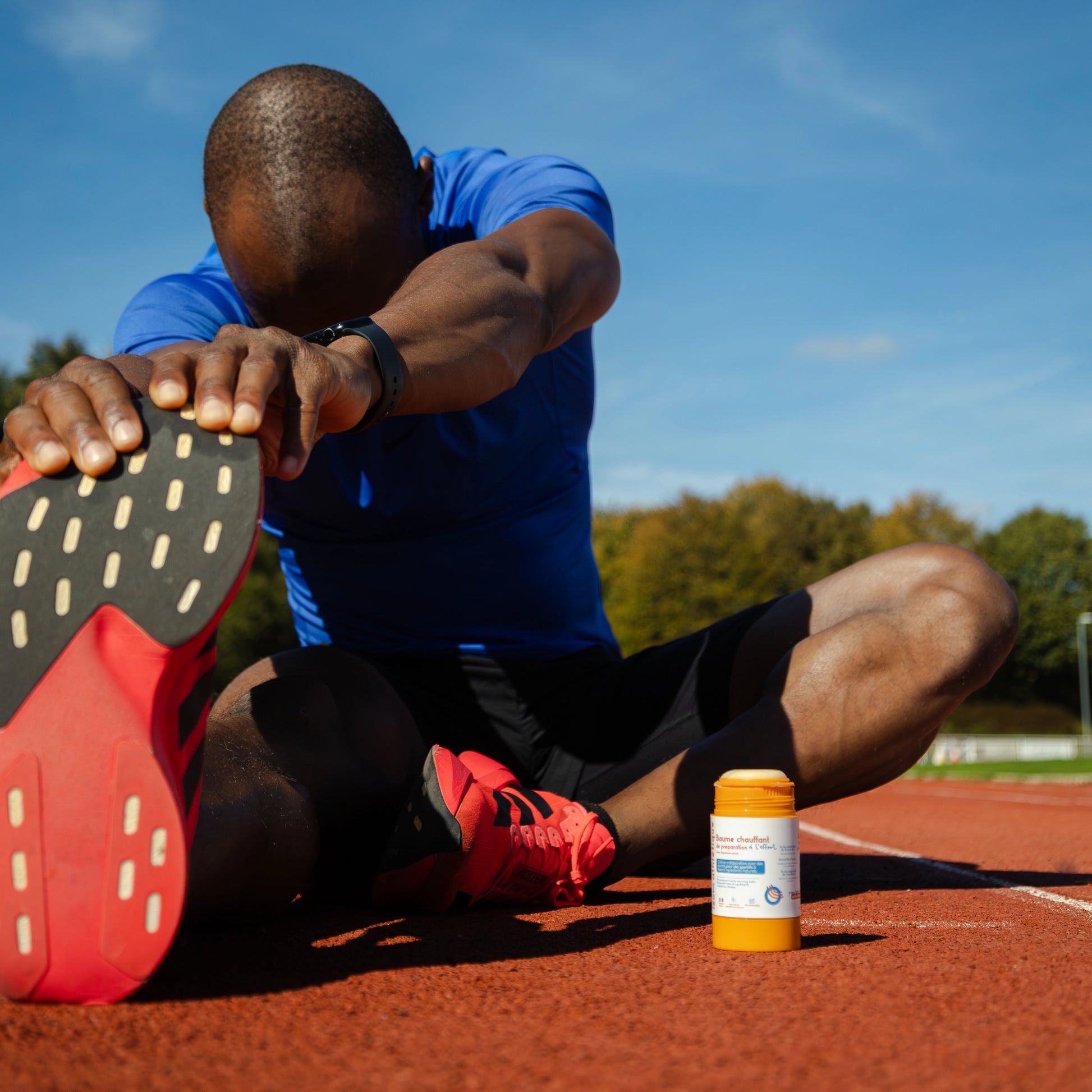 Oumar BAMBA, athlète (sprinteur) et ambassadeur IZOM Athlétique s'étire les muscles sur une piste d'athlétisme. Le baume chauffant muscles 100% naturel et rechargeable IZOM Athlétique est posé ouvert à côté de lui. La texture baume est visible.